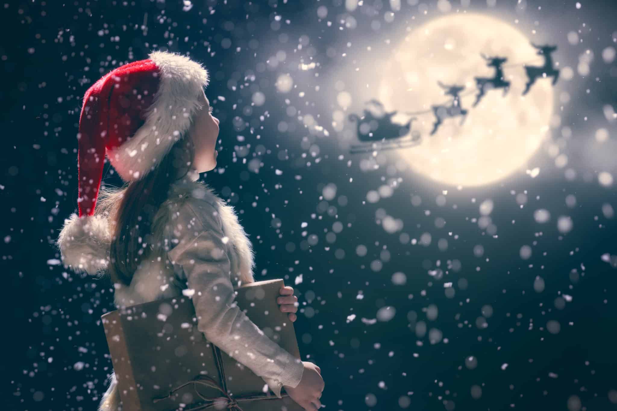 Girl with Christmas hat looking at the shadow of Santa and his reindeers passing in front of the moon