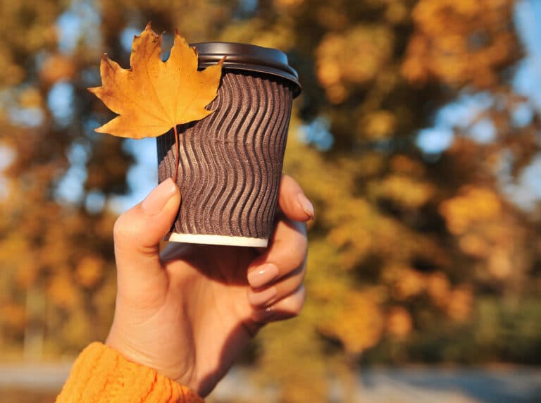 Hand holding a takeaway coffee cup with an autumn leaf in front of golden trees at a dog-friendly café near Bewl Water.
